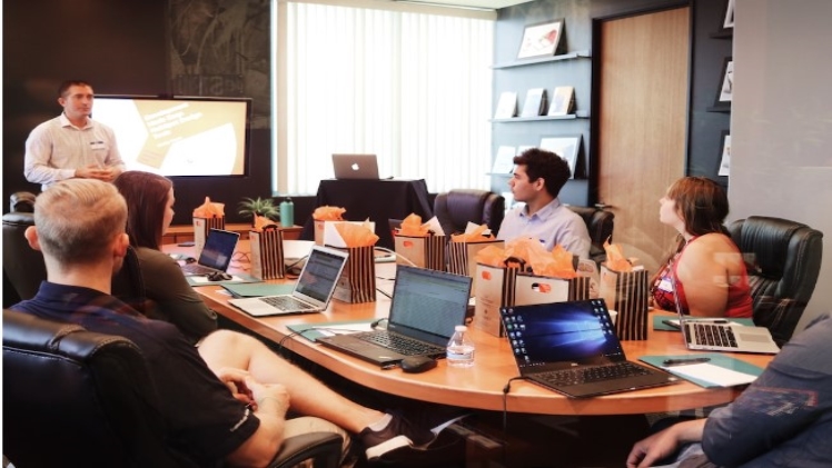 A manager discusses capacity utilization in front of a team sitting at a table with laptops.